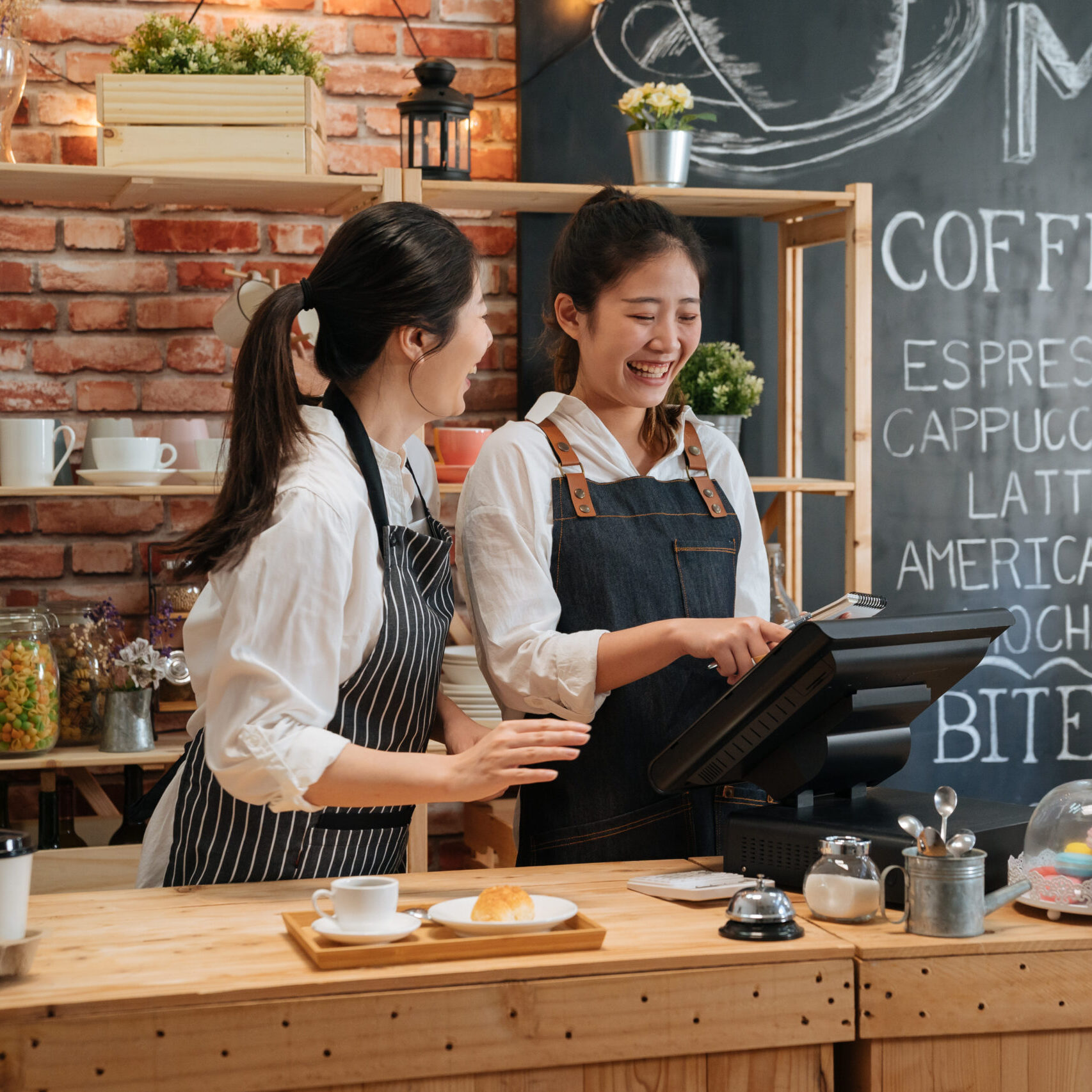 asian female barista telling something funny to colleague while working. two young girls coffeehouse staff laughing together and chatting gossip in modern coffee store. customer meal on bar counter