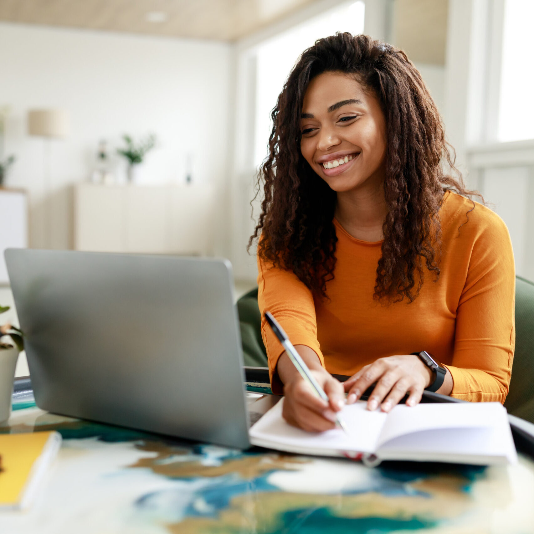 Smiling young African American woman sitting at desk working on laptop taking notes in notebook, happy millennial female studying online, watching webinar using computer and writing check list